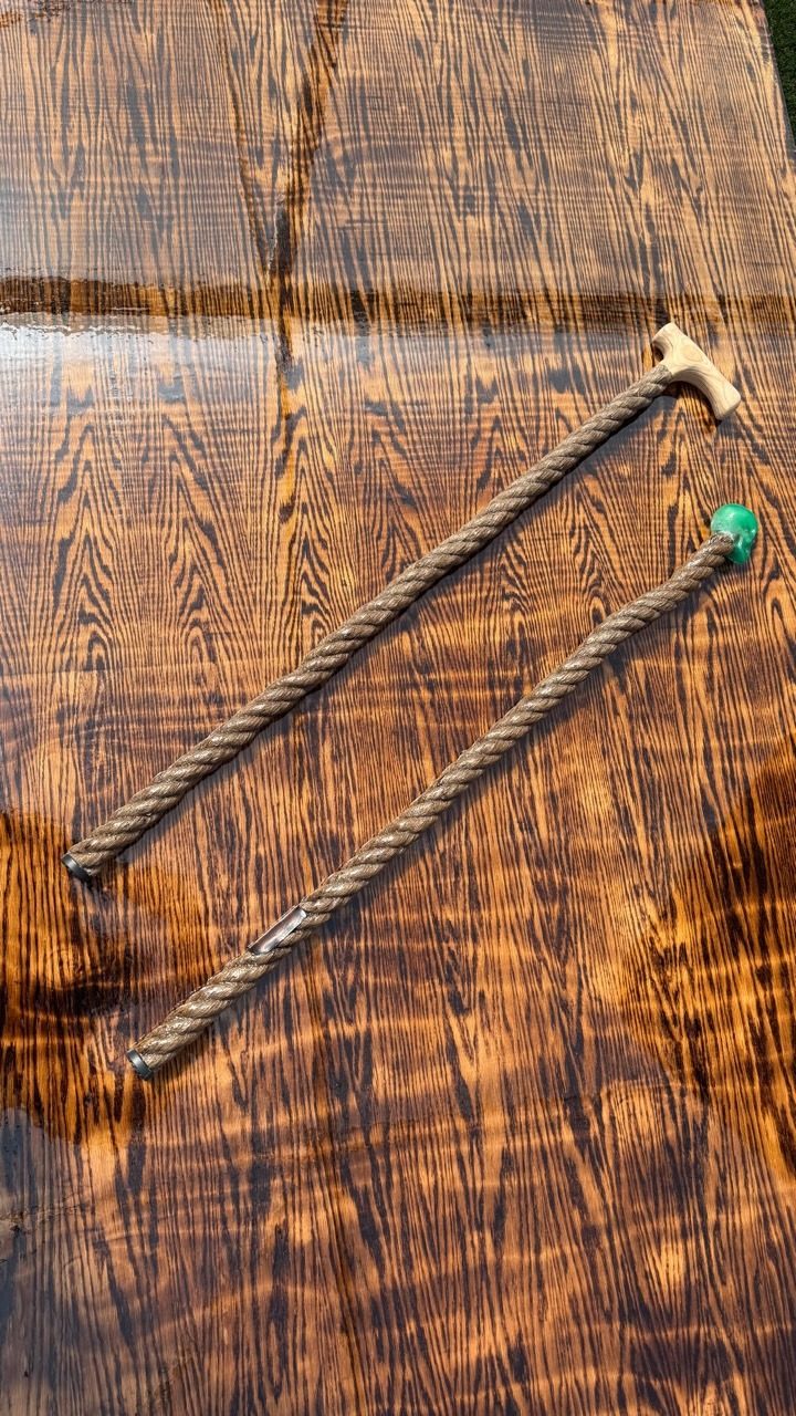 Two handmade walking pieces on a dark wood surface, one with a carved rope shaft and pale T handle, the other with a green skull finial.