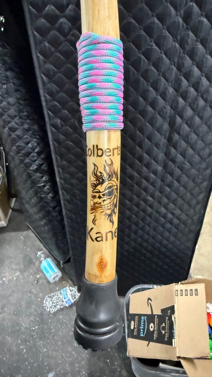 A light-wood handled cane with a multicolor paracord wrap, woodburned mark, and black rubber foot.